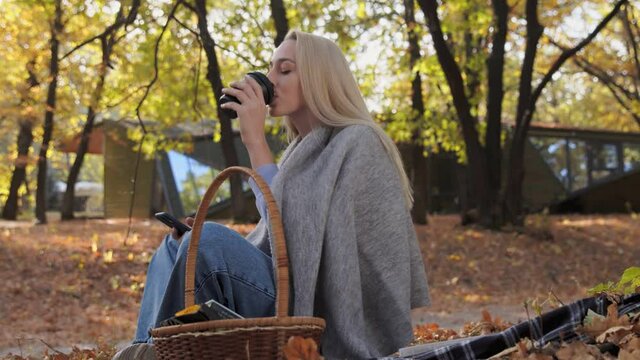 Young blonde woman drinks coffee and uses phone in the park at fall on a sunny day