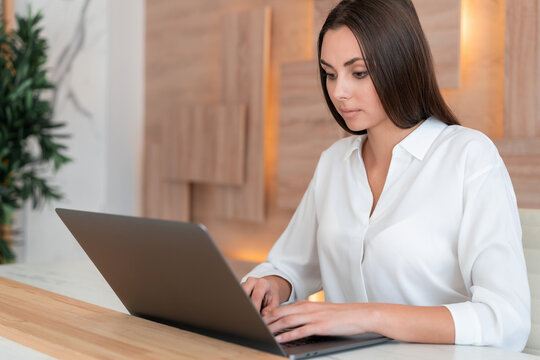 Office manager woman wearing white shirt, sitting to table with laptop on background of wooden office wall, side view. Concentrated office worker looking at the screen. Concept of work