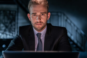 Portrait of tired businessman wearing black suit, working late at night in the office. Office manager working with laptop looking at the camera, blurred background of dark office room