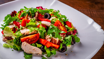 Fresh salad with chicken breast, arugula, black olives,red pepper, lettuce, fresh sald leaves and tomato on a white plate on wooden table background