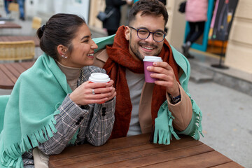 A young couple below the blanket is sitting and drinking coffee outside in the coffee bar.