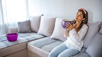 Obraz premium Frustrated homeowner watching ceiling leaks sitting on a couch at home. Worried Woman Holding Two Buckets While Water Droplets Leaking From Ceiling.