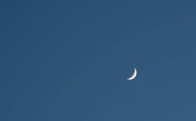 A slim sliver of the silvery moon set on an expanse of a partly cloudy blue sky.
