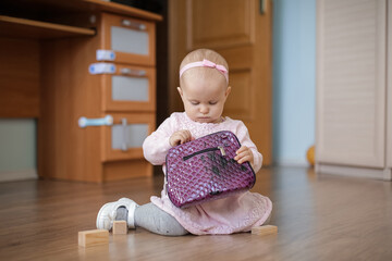 A one-year-old baby in a cute pink dress with a tulle skirt sits on the floor with a purple bag.