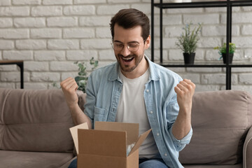Emotional happy young man in eyeglasses opening small cardboard parcel, celebrating getting wished...