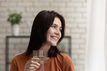Happy millennial attractive woman holding glass of fresh mineral water, looking away, daydreaming...