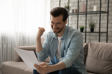 Emotional happy positive young man in eyewear reading last mortgage payment paper bank...