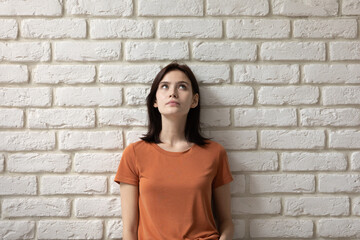 Fototapeta na wymiar Dreamy young caucasian woman looking up in distance isolated on white brick wall background, pensive millennial lady thinking of problem solution ideas, planning or visualizing future, copy space.