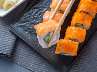Philadelphia and California rolls on a textured dark plate. One roll is held in Granny sticks above the others. Next to the wasabi and ginger. Side view. Japanese cuisine