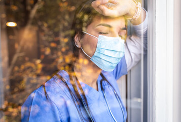 Portrait of exhausted nurse with mask standing behind the glass.