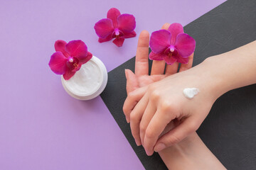 Cosmetic background with hands, soft white cream and fragile magenta colored orchids.Moisturizing facial cream in a jar and blooming orchid flowers on purple and black background, skin care cosmetics.