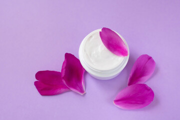 Container with white moisturizing cream with peony extract and petals on pink background