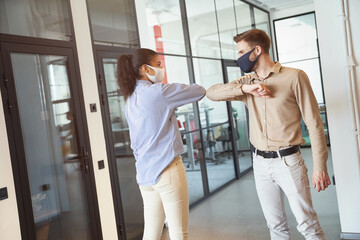 Covid 19 infection prevention. Two young diverse colleagues wearing face protective masks bumping elbows, greeting each other while standing in the modern office