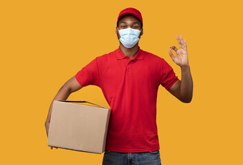 Fototapeta na wymiar Black delivery man in mask holding box, showing okay gesture