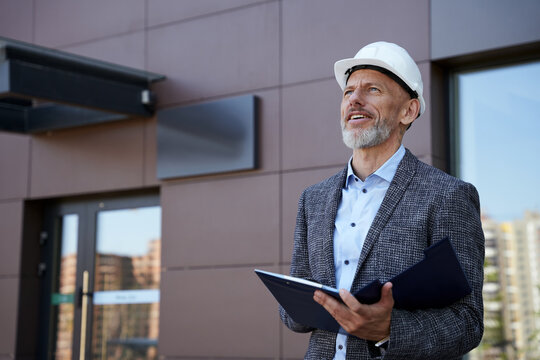 Monitoring The Process. Confident Middle Aged Architect, Engineer Manager Wearing Helmet Looking Away While Standing Outdoors And Holding A Blueprint