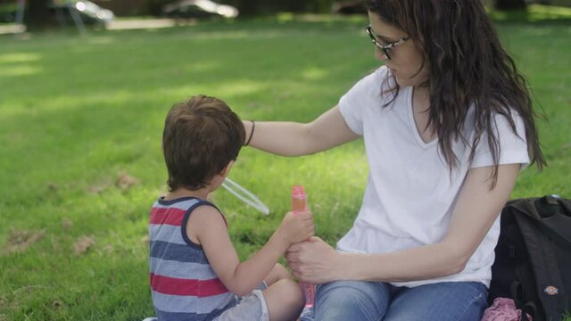 A beautiful mom and her son blow bubbles while sitting a picnic blanket in a park