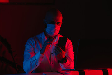 A bald man in a medical face mask is working remotely on a laptop at home. A guy with earphones is scrolling the news on a smartphone. A male employee in front of the computer under blue and red light