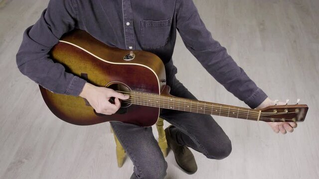 A musician tunes an acoustic guitar. Guitar player holds brown vintage acoustic guitar and pulls strings. He wears black denim jeans and shirt, brown shoos. Guitar player school. White background. 4k