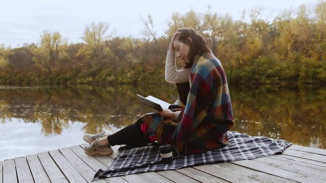 Young beautiful woman wint blanket on shoulders reads book and drinks coffee while stits po piea by the river