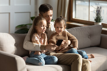 Daddy hug little daughters sit on couch in living room with smartphone. Father teach show to kids how to use device, educate children watch together development cartoons, have fun, play games concept