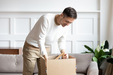 Obraz na płótnie Canvas Man standing in living room on day of move to rented home, unbox carton box with belongings. Relocate to new own apartment collect personal things. Delivered parcel to satisfied client, tenant concept