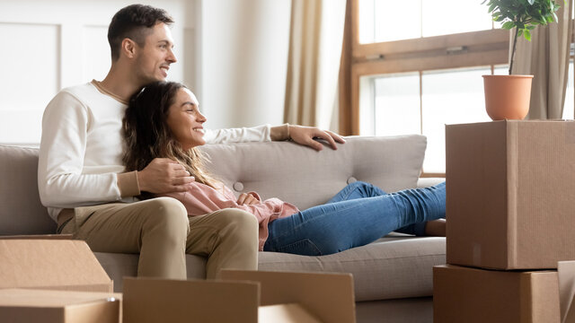 Couple resting on couch on moving day to new apartment, boxes with packed stuff on floor, spouses dreaming about happy future in own home, plan rooms interior design. Bank loan, first realty concept