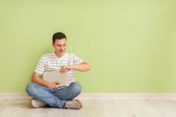 Young man with wristwatch and laptop sitting near color wall