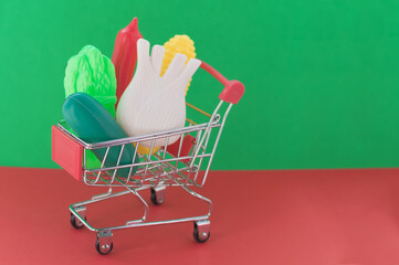 Shopping cart on a red and green background with plastic vegetables inside. The concept of non-natural products with harmful additives.