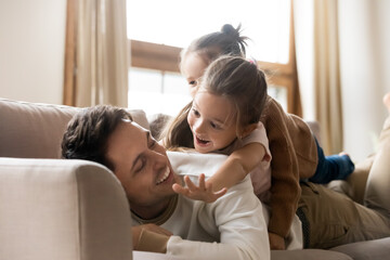 Fototapeta premium Two cheerful preschool daughters lying on father back spend funny weekend time together indoor, communicating enjoy relaxation and playtime at home. Family bond, love to kids, happy fatherhood concept