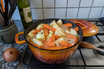 Orange casserole with meat, sliced potatoes, carrots and celeriac on gas stove. Onion studded with cloves. Cooking scenery for concept.