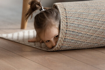 Adorable small girl wrapped in rug lying on warm wooden laminate floor in modern living room alone,...