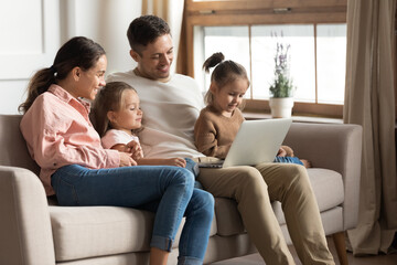Beautiful full family with two cute daughters resting with laptop seated on sofa. Couple develop...