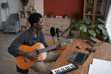 High angle portrait of young African-American man playing guitar and singing to microphone in home...