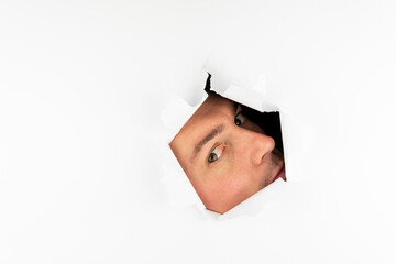 A man looks through a hole in a white wall with one eye.
