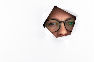 A woman with black-rimmed glasses looks through a hole in a white paper wall. Free space for text.
