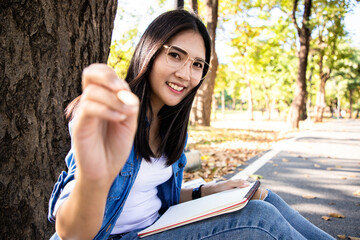 Beautiful asian woman holding pencil and writing diary on notebook with bright smile face in public park. She learning and work hard in the last weekend. Spring season warm tone
