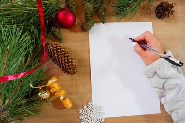 A girl writes a New Year's greeting card or Christmas wish sheet on a white piece of paper. Background of Christmas tree toys and evergreen pine needles