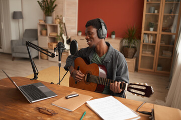 Portrait of smiling African-American man singing to microphone and playing guitar while recording...