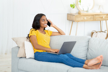 Joyful African American woman communicating with someone on cellphone and laptop from home