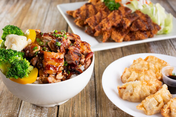 A view of several pan asian entrees prepared on a wooden table surface, featuring a teriyaki chicken bowl, potstickers, and chicken katsu.