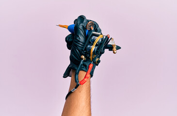 Hand of young hispanic man using tattoo machine over isolated pink background.