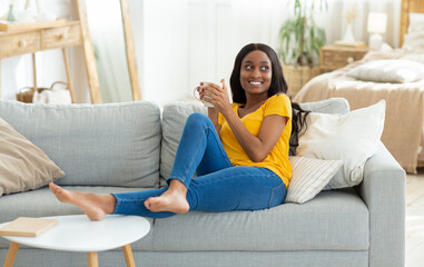 Lovely young black woman having coffee break on comfy couch at home, copy space