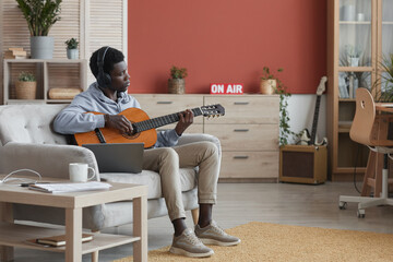 Full length portrait of young African-American man playing acoustic guitar while sitting on couch...