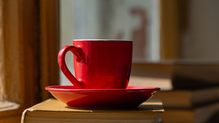 red cup with coffee stands on old books against the background of the window.