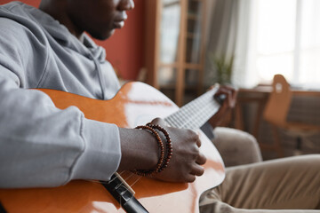 Close up of young African-American man playing acoustic guitar while sitting on couch and making music at home, copy space