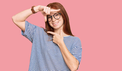 Young read head woman wearing casual clothes and glasses smiling making frame with hands and...