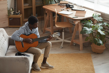 Full length portrait of young African-American man playing acoustic guitar while sitting on couch...