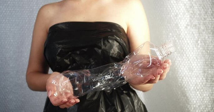 Plastic pollution. Ecology problem. Waste reduction. Female volunteer in black garbage bag dress holding crumpled used disposable bottle in hands on blur bubble wrap background.