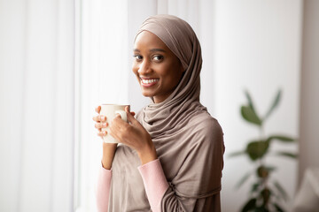 Black Muslim woman in traditional hijab drinking hot aromatic coffee near window at home, having...