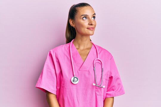Young hispanic woman wearing doctor uniform and stethoscope smiling looking to the side and staring away thinking.